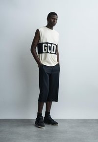 Un homme est appuyé contre un mur uni, portant un haut blanc sans manches avec le texte "GCDS", un pantalon noir large coupé, des chaussettes noires et des baskets noires.