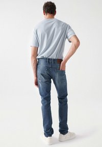 T-shirt azzurro abbinato a jeans blu scuro con due tasche posteriori e una toppa in pelle con marchio. Sneakers bianche completano l'outfit.