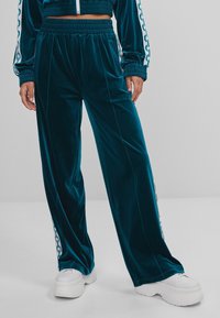 Tyrkysové sametové kalhoty s širokými nohavicemi, zdobené bočními pruhy a pohodlným elastickým pasem. Kombinované s robustními bílými teniskami.