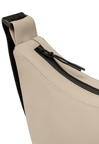 Beige keresztpántos táska, sima, matt felülettel, fekete cipzárral és húzófüllel, valamint kontrasztos fehér varrással a széleken.