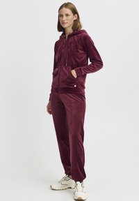 Survêtement en velours bordeaux avec une veste à capuche zippée et des poches, associé à des chaussures de sport. Texture douce et coupe décontractée.
