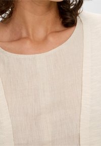 Béžový lněný top s kulatým výstřihem a texturovaným kardiganem s otevřeným předním dílem. Tkanina je lehká a mírně průsvitná, s minimálními detaily švů.