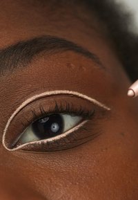 Närbild av ett öga med mörkbrun hy, med en ljus beige eyeliner applicerad längs den övre franslinjen, vilket framhäver ögats form och detaljer.