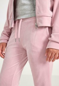 Lyserøde velour joggers med en glat tekstur, der har en snor i taljen og sølvfarvede hardware-detaljer. Stylet med en cropped grå top.