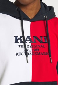 Felpa con cappuccio a pannelli rossi, bianchi e blu navy. Presenta un logo ricamato con la scritta "KANI THE ORIGINAL EST. 1989" in blu navy. Tessuto in misto cotone.
