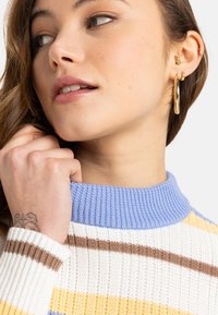 Strickpullover mit blauen, gelben und braunen Streifen. Rippkragen und -bündchen. Das Model hat ein Ohrpiercing und ein Blatttattoo auf ihrer Hand.
