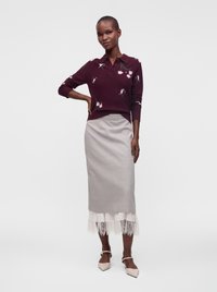 Femme portant un haut à manches longues bordeaux à motif floral, une jupe midi grise avec un bord en dentelle blanche et des chaussures pointues beiges, posant avec une main sur la hanche.