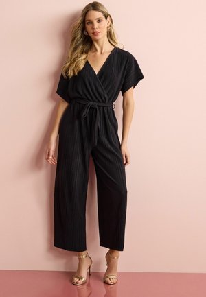 Černý jumpsuit s pruhy a překládaným designem, krátkými rukávy a zavázaným pasem. V kombinaci se zlatými sandály na vysokém podpatku se zavazováním.