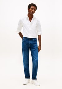 Mladý muž stojící s rukama za zády, oblečený v bílé košili na knoflíky, modrých džínách s ohrnutými lemy a bílých teniskách na jednobarevném pozadí.