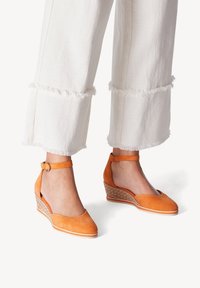 Sandales à talons compensés en suede orange avec un bout rond, une sangle à la cheville et un talon texturé tissé. Associées à un pantalon large à ourlet effiloché blanc.