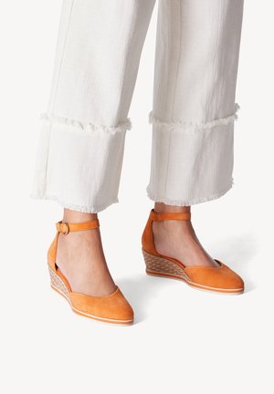 Orange suede wedge sandals with a round toe, ankle strap, and woven textured heel. Paired with white, frayed hem wide-leg pants.