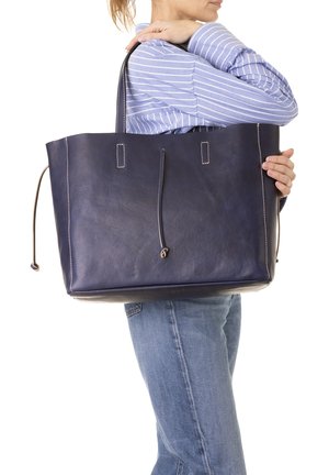 Femme en chemise rayée et jean, tenant un grand sac cabas en cuir bleu marine sur l'épaule, devant un fond blanc.