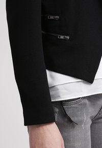 Černý tailored blazer s hladkou texturou, se zipovými kapsami a asymetrickým spodním lemem, nošený přes bílou vrstvenou blůzu a šedé džíny.