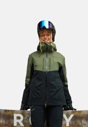 Frau in grüner und schwarzer Winterjacke, die Skibrille trägt und ein Snowboard mit braun-weißem Design hält, lächelnd mit geschlossenen Augen.