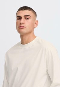 Camisa de manga larga beige claro con cuello redondo, que presenta una textura suave y lisa y un corte relajado. Diseño simple sin patrones visibles.