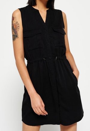 Robe noire sans manches avec poches poitrine et taille à cordon, portée par une personne avec un tatouage sur le haut du bras et la main dans la poche.