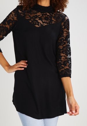 Tunique noire avec dentelle florale sur le haut de la poitrine et manches trois-quarts, portée avec un jean bleu clair sur une personne debout.