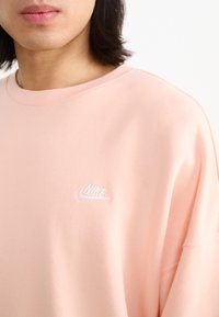 Rožinė Nike firminė palaidinė su apykakle, ilgomis rankovėmis ir siuvinėtu baltu logotipu ant krūtinės. Minkštas, glotnus audinys ir laisvas kirpimas.
