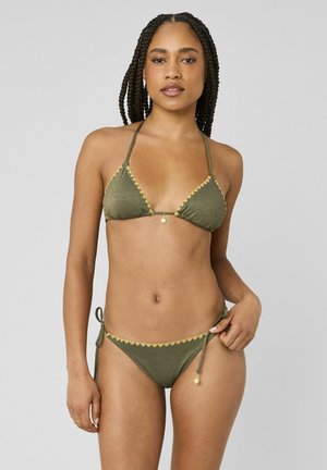 Femme aux cheveux tressés portant un bikini métallique vert avec un bord festonné jaune et de petits pendentifs en forme de soleil, debout devant un fond gris uni.