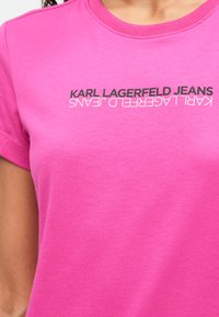 Tricou roz cu mânecă scurtă, fabricat din bumbac moale, având textul "KARL LAGERFELD JEANS" tipărit cu litere mari negre pe piept.