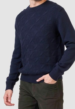 Homme portant un pull en tricot texturé bleu marine et un pantalon foncé, avec une main dans la poche, devant un fond clair et uni.