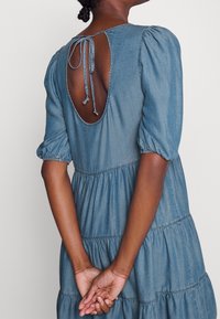 Vestido de mezclilla en azul claro con mangas abullonadas, diseño en capas y un escote abierto atado con cuerdas a juego. Tejido suave y texturizado.