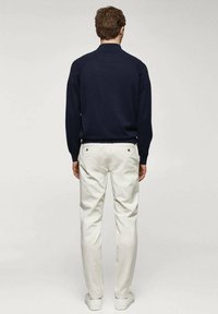 Un pull bleu marine avec un col haut, associé à un pantalon couleur crème. Le pantalon comporte des poches arrière avec des boutons métalliques ronds.