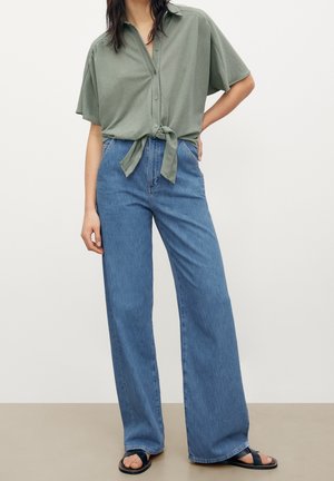 Femme portant une chemise ample vert clair à manches courtes boutonnée et nouée à la taille, un jean bleu à jambes larges et des sandales noires sur fond uni.
