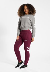 Grauer Pullover mit "HUMMEL"-Logo, kombiniert mit burgunderfarbenen Jogginghosen mit weißen Chevron-Akzenten und Taschen. Schwarze Sportschuhe runden das Outfit ab.