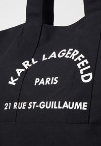 Black tote bag made of durable fabric featuring bold white text: "KARL LAGERFELD PARIS 21 RUE ST-GUILLAUME." Simple design, straight handles.