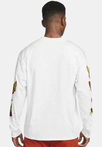 Långärmad vit skjorta med färgglada fjärilsdesign på båda ärmarna, med rundad halsringning och en avslappnad passform.