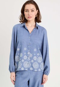 Chemise bleu à boutons, fabriquée en tissu léger. Elle possède des manches longues, un col et une broderie florale blanche à l'ourlet. Texture douce.