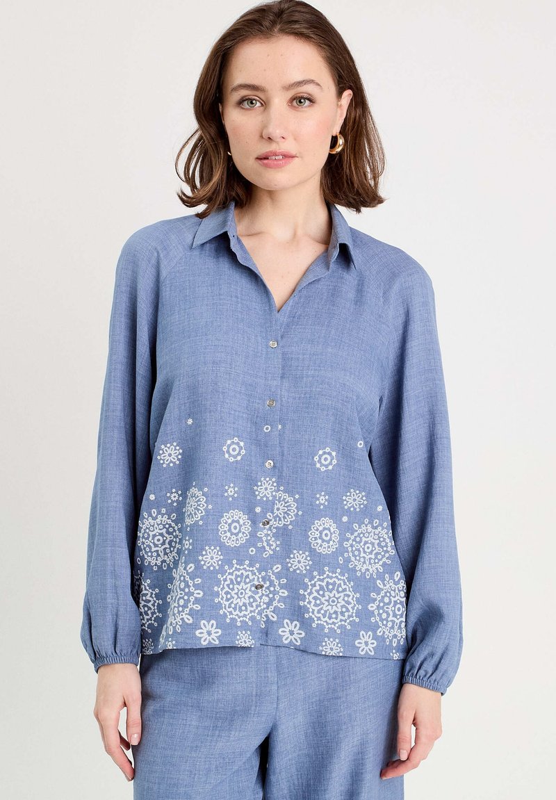 Chemise bleu à boutons, fabriquée en tissu léger. Elle possède des manches longues, un col et une broderie florale blanche à l'ourlet. Texture douce.