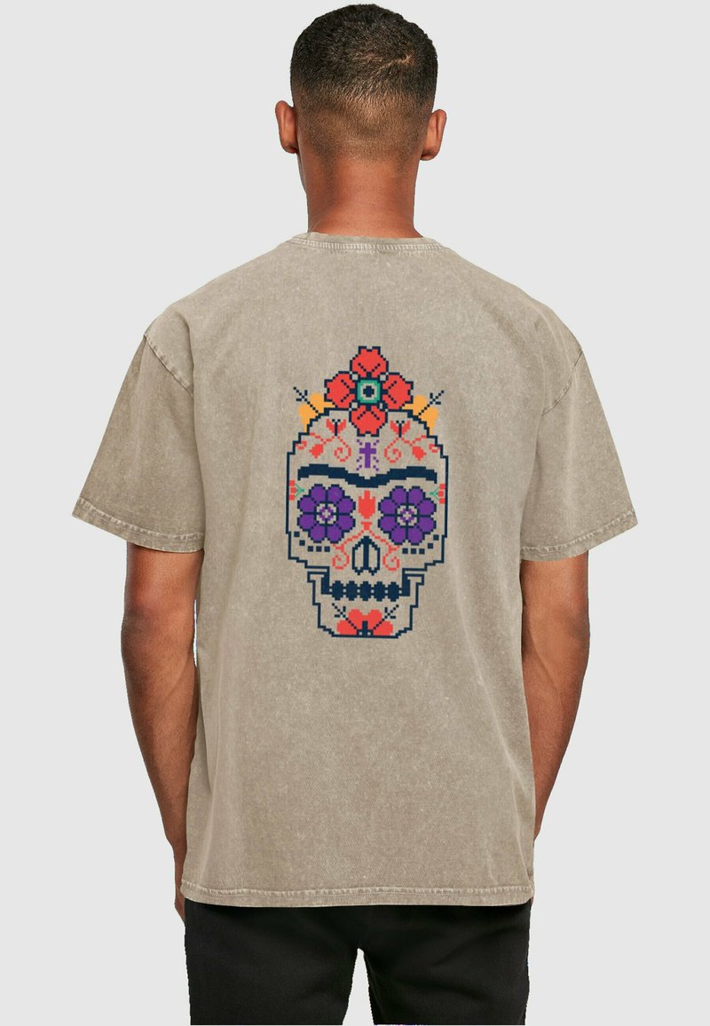 Lyse beige kortærmet t-shirt med et farverigt, tegneserieagtigt kraniumgrafik på ryggen, prydet med blomster og dekorative detaljer.