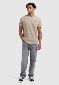 Beige katoenen t-shirt met korte mouwen en een ronde halslijn, met een klein zwart logo. Gekozen in combinatie met grijze denim jeans en zwarte sneakers.