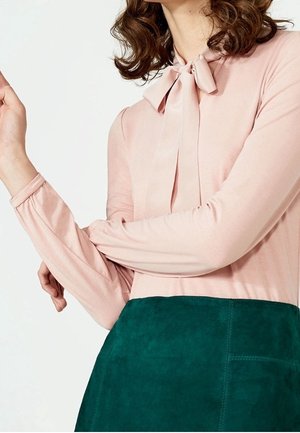 Nainen pukeutuneena vaaleanpunaiseen, pitkähihappiseen paitaan, jossa on rusetti kauluksessa, ja tummanvihreään mokkahameeseen, poseeraa käsivarret ristissä.