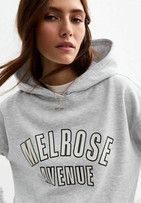 Gray hoodie featuring "Melrose Avenue" in bold black letters, a kangaroo pocket, and a drawstring hood. Soft, cotton blend texture.