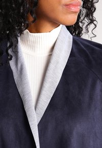 Marinblå mockajacka med en ljusgrå stickad krage, med en sjalkrage-design. Texturerat tyg, öppen framtill och utan synliga knäppningar.