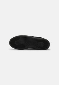 Suela de zapatilla negra con una superficie texturizada, un patrón de banda de rodadura distintivo y un detalle de logo redondo. Material de goma duradero con un diseño plano.