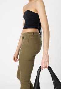 Top negro sin tirantes con textura fruncida; vaqueros ajustados verde oliva; persona sosteniendo un bolso de cuero negro con asa curvada.