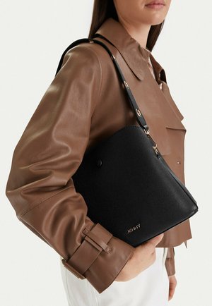 Kobieta w brązowej skórzanej kurtce trzyma czarną torbę na ramię o fakturze, z złotymi elementami i logo "JENNY".