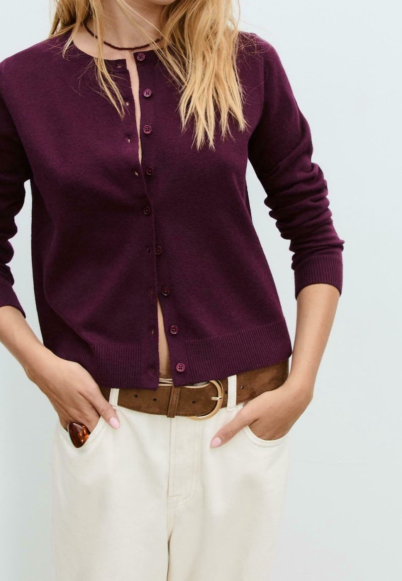 Femme portant un cardigan violet foncé boutonné, pantalon beige avec une ceinture marron, les mains dans les poches, et portant une bague à la main gauche.