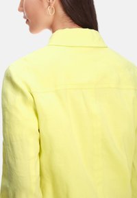 Light yellow collared jacket with a textured fabric. Features visible stitching and a straight back design. Minimalist and modern style.