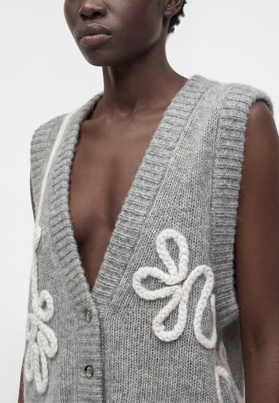 Personne noire portant un gilet sans manches gris en maille avec un col en V profond et de grands motifs floraux blancs tricotés à l’avant et sur les côtés.