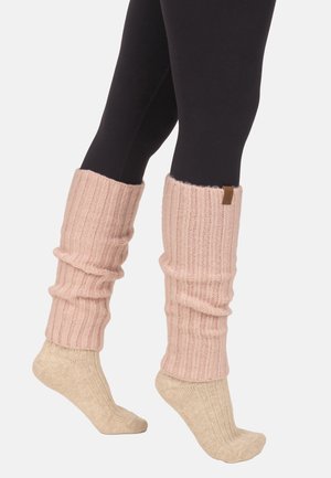 Nogi w czarnych legginsach, na których założone są beżowe prążkowane skarpetki i różowe dziergane ocieplacze na nogi, stojące na palcach na białym tle.