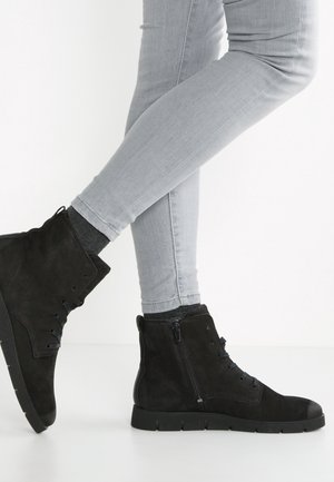 Une personne portant un jean gris clair et des bottines noires à lacets avec des fermetures éclair latérales se tient sur un fond blanc, un pied levé.
