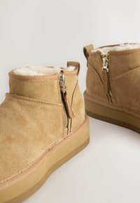 Sandy ruskindankelstøvler med lynlås, faux fur-foring og en flad, struktureret gummisål. Har synlige sømme og en løkke.