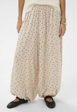 Beige Hosen mit weitem Bein und schwarzen Punkten, elastische Bündchen am Knöchel, kombiniert mit schwarzen Slippern und einem goldenen Armband am rechten Handgelenk.