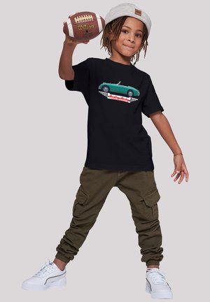 Schwarzes T-Shirt mit einem bunten Vintage-Auto-Graphic, olivgrüne Cargo-Hosen und weiße Sneakers. Das Kind hält einen braunen Football und trägt eine hellgraue Mütze.