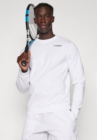 Sweatshirt blanc à manches longues avec un col rond et le logo Kappa sur la poitrine gauche, tenu par une personne avec une raquette de tennis sur un fond uni.